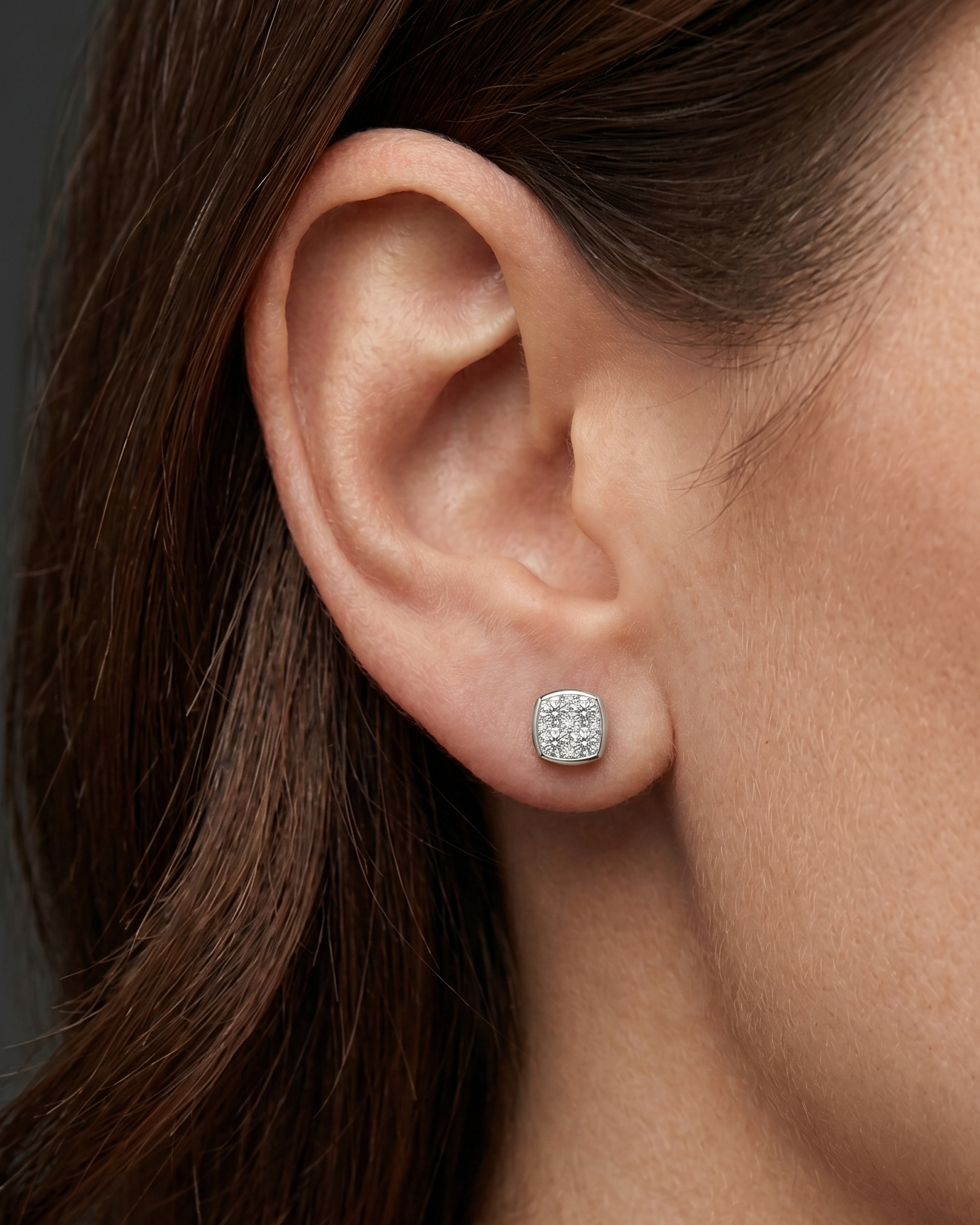 A close-up, editorial-style photograph focusing on a woman's ear and jawline to showcase a petite, cushion-shaped stud earring. The silver-toned earring features a cluster of sparkling clear gemstones set within a smooth metallic border. The model has dark brown hair tucked behind her ear, set against a seamless dark charcoal gray background.