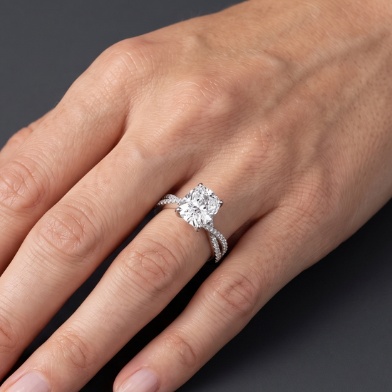 A close-up of a woman's hand wearing a sparkling engagement ring with an elongated cushion-cut center diamond and a twisted pavé band, resting gracefully against a smooth, dark charcoal gray background.