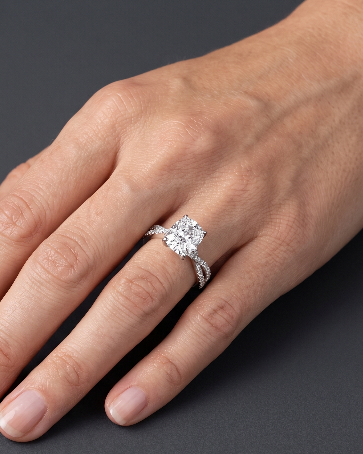 A close-up of a woman's hand wearing a sparkling engagement ring with an elongated cushion-cut center diamond and a twisted pavé band, resting gracefully against a smooth, dark charcoal gray background.