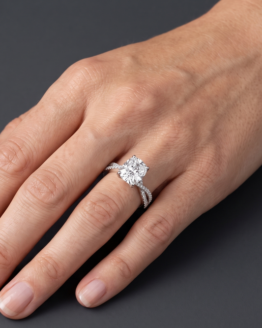A close-up of a woman's hand wearing a sparkling engagement ring with an elongated cushion-cut center diamond and a twisted pavé band, resting gracefully against a smooth, dark charcoal gray background.